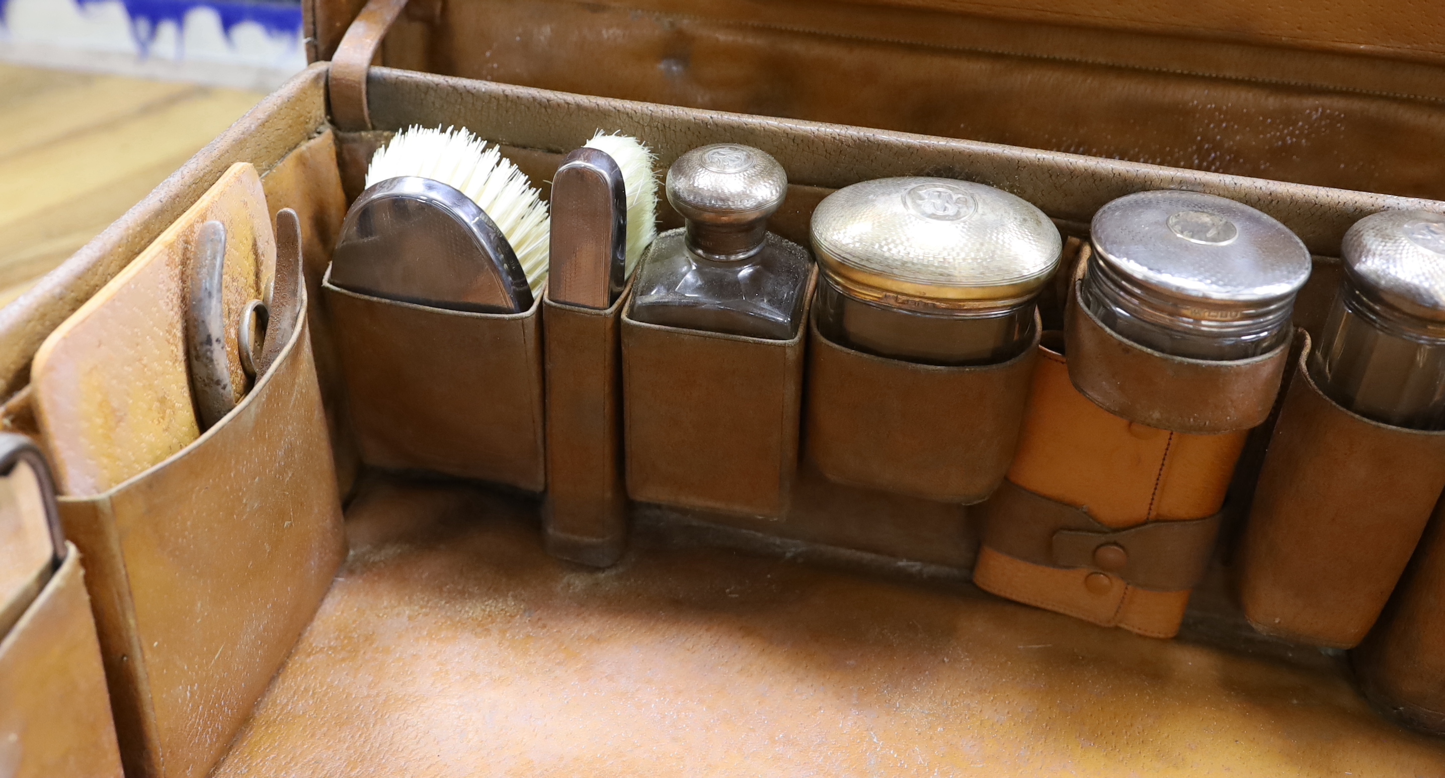 An Edwardian leather travelling toilet case, containing eleven silver mounted toilet jars etc. including a mounted glass hip flask and easel mirror, Drew & Sons, London, 1905, case width 62.5cm.